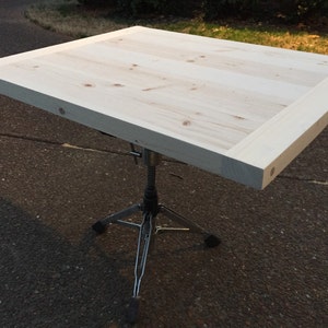 May include: A light-colored wooden table, approximately 30 inches square, with a natural wood grain finish. The tabletop is supported by a silver metal tripod base, creating a simple, modern aesthetic.