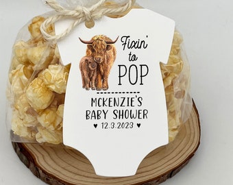 May include: A white baby onesie favor tag with a brown Highland cow and calf illustration. The tag says "Fixin' to POP McKenzie's Baby Shower 12.3.2023".