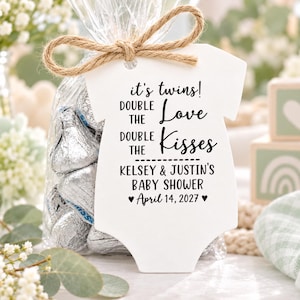 May include: A white onesie-shaped tag tied with a jute twine bow to a clear plastic bag filled with silver-wrapped chocolate candies. The tag features black text reading: it's twins! DOUBLE THE Love DOUBLE THE Kisses KELSEY & JUSTIN'S BABY SHOWER &hearts; April 14, 2027 &hearts;.
