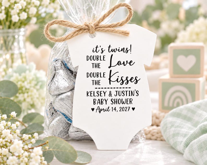 May include: A white onesie-shaped tag tied with a jute twine bow to a clear plastic bag filled with silver-wrapped chocolate candies. The tag features black text reading: it's twins! DOUBLE THE Love DOUBLE THE Kisses KELSEY & JUSTIN'S BABY SHOWER &hearts; April 14, 2027 &hearts;.