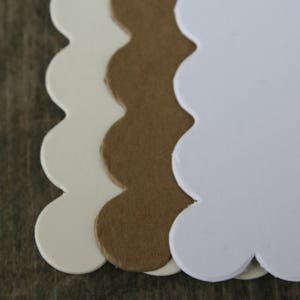 May include: Three scalloped-edge paper tags or labels arranged in a vertical stack. The top layer is bright white, the middle layer is a natural brown kraft paper, and the bottom layer is a light cream color. Each piece has a smooth, rounded edge.