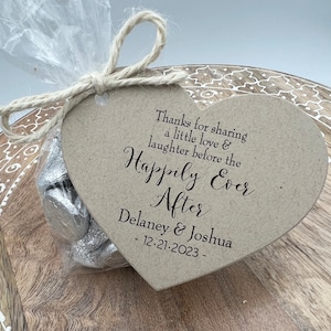 Puede incluir: Una etiqueta marr&oacute;n en forma de coraz&oacute;n con el texto "Thanks for sharing a little love & laughter before the Happily Ever After Delaney & Joshua - 12.21.2023 -" impreso en ella. La etiqueta est&aacute; atada con un cordel a una bolsa de pl&aacute;stico transparente llena de chocolates envueltos en papel plateado.