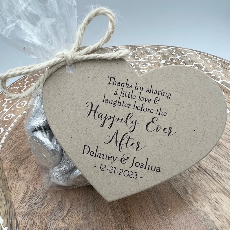 Puede incluir: Una etiqueta marr&oacute;n en forma de coraz&oacute;n con el texto "Thanks for sharing a little love & laughter before the Happily Ever After Delaney & Joshua - 12.21.2023 -" impreso en ella. La etiqueta est&aacute; atada con un cordel a una bolsa de pl&aacute;stico transparente llena de chocolates envueltos en papel plateado.