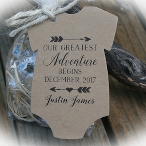 May include: A kraft paper onesie-shaped tag with the text "Our Greatest Adventure Begins December 2017 Justin James" in black script. The tag is tied to a small bag with a twine bow. The background is a rustic wooden surface.