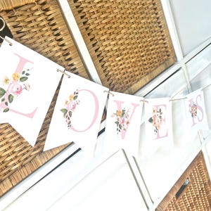 May include: A white banner with pink letters spelling "LOVE IS" decorated with floral designs. The banner is strung with twine and hung on a woven wicker surface.