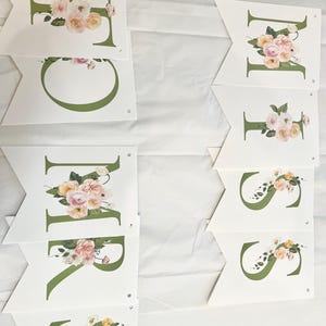 May include: A floral banner with the word "MRS" spelled out in green letters with floral accents. The letters are on white flags with holes for hanging.