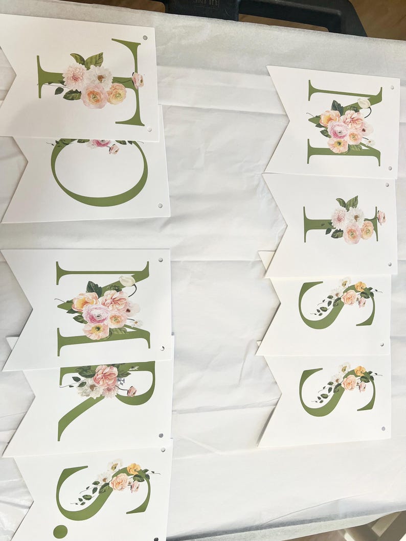 May include: A floral banner with the word "MRS" spelled out in green letters with floral accents. The letters are on white flags with holes for hanging.