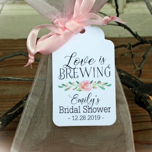 以下が含まれることがあります： ピンクのリボンと「Love is Brewing Emily's Bridal Shower - 12.28.2019 - 」と書かれたタグが付いた白いギフトバッグ。タグには、ピンクの花と緑の葉の入った花柄のデザインが施されています。