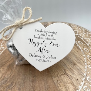 Puede incluir: Etiqueta de madera blanca en forma de coraz&oacute;n con el texto "Thanks for sharing a little love & laughter before the Happily Ever After Delaney & Joshua - 12-21-2023 -" impreso en negro. La etiqueta est&aacute; atada con un cordel a una bolsa de celof&aacute;n transparente llena de corazones de chocolate envueltos en papel de aluminio plateado.