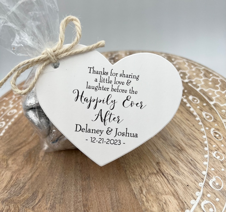 Puede incluir: Etiqueta de madera blanca en forma de coraz&oacute;n con el texto "Thanks for sharing a little love & laughter before the Happily Ever After Delaney & Joshua - 12-21-2023 -" impreso en negro. La etiqueta est&aacute; atada con un cordel a una bolsa de celof&aacute;n transparente llena de corazones de chocolate envueltos en papel de aluminio plateado.