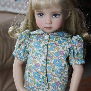 May include: A porcelain doll with blonde hair wearing a white and blue floral print dress with a white collar and buttons. The doll has blue eyes and rosy cheeks.
