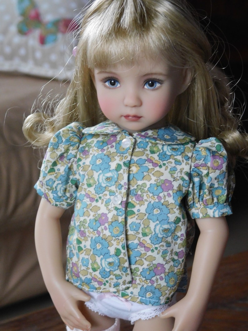 May include: A porcelain doll with blonde hair wearing a white and blue floral print dress with a white collar and buttons. The doll has blue eyes and rosy cheeks.