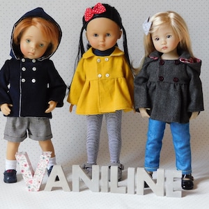 Peut inclure: Trois poupées portant des manteaux de couleurs différentes. La poupée à gauche porte un manteau bleu marine avec des boutons blancs et des bordures blanches. La poupée du milieu porte un manteau jaune avec des boutons blancs. La poupée à droite porte un manteau gris avec des bordures rouges. Les poupées sont debout devant le mot "VANILINE".