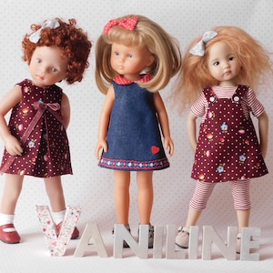 May include: Three dolls wearing different outfits. The doll on the left is wearing a burgundy dress with white polka dots and a white bow. The doll in the middle is wearing a blue denim dress with a red heart and white trim. The doll on the right is wearing a burgundy dress with white polka dots and a red and white striped shirt. The dolls are standing in front of the word "VANILINE".