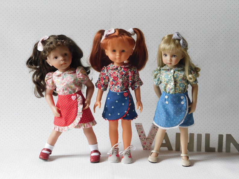 May include: Three dolls wearing floral print shirts and different colored skirts. The doll on the left is wearing a red skirt with white polka dots and a white ruffle trim. The doll in the middle is wearing a blue denim skirt with white hearts and a white ruffle trim. The doll on the right is wearing a blue denim skirt with white polka dots and a white lace trim.