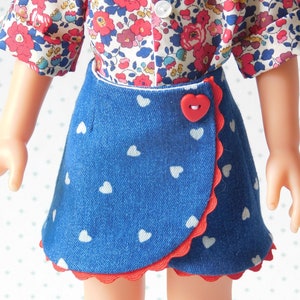 May include: A blue denim skirt with white heart print and red trim. The skirt has a wrap-around style and a red heart-shaped button.