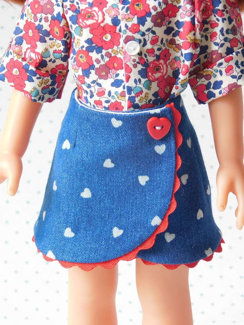 May include: A blue denim skirt with white heart print and red trim. The skirt has a wrap-around style and a red heart-shaped button.