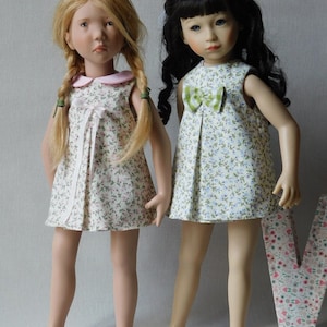 Puede incluir: Dos muñecas con vestidos de estampado floral. La muñeca de la izquierda tiene el pelo rubio y un cuello rosa en su vestido. La muñeca de la derecha tiene el pelo negro y un lazo verde y blanco en su vestido.