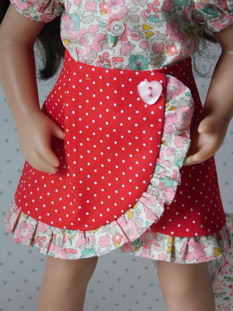 May include: A red skirt with white polka dots and a floral ruffle trim. The skirt is wrapped and has a pink heart-shaped button.