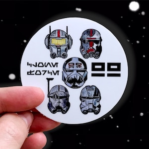 May include: A white round sticker with five black and white illustrations of Star Wars clone trooper helmets. The text "BAD BATCH" is printed in black letters on the sticker.