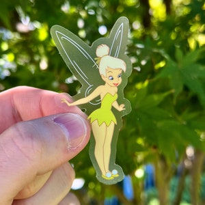 K&ouml;nnte beinhalten: Ein transparenter Aufkleber mit Tinkerbell, einer Disney-Fee mit blonden Haaren, gr&uuml;nen Fl&uuml;geln und einem gelben Kleid. Sie steht mit ausgestreckten Armen und einem L&auml;cheln im Gesicht.