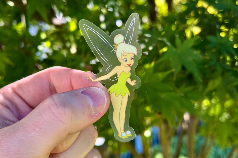 K&ouml;nnte beinhalten: Ein transparenter Aufkleber mit Tinkerbell, einer Disney-Fee mit blonden Haaren, gr&uuml;nen Fl&uuml;geln und einem gelben Kleid. Sie steht mit ausgestreckten Armen und einem L&auml;cheln im Gesicht.