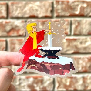 Pode incluir: Um adesivo com um personagem de desenho animado puxando uma espada de uma pedra. O personagem est&aacute; vestindo uma t&uacute;nica vermelha e um cachecol amarelo. O fundo &eacute; uma paisagem nevada com flocos de neve.