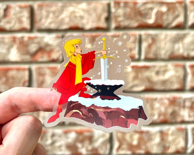 Pode incluir: Um adesivo com um personagem de desenho animado puxando uma espada de uma pedra. O personagem est&aacute; vestindo uma t&uacute;nica vermelha e um cachecol amarelo. O fundo &eacute; uma paisagem nevada com flocos de neve.