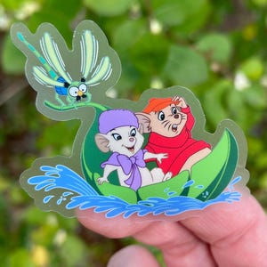 May include: A clear sticker featuring two cartoon mice in a green leaf boat, with a dragonfly flying above them. The mice are wearing hats and the boat is floating on blue water.