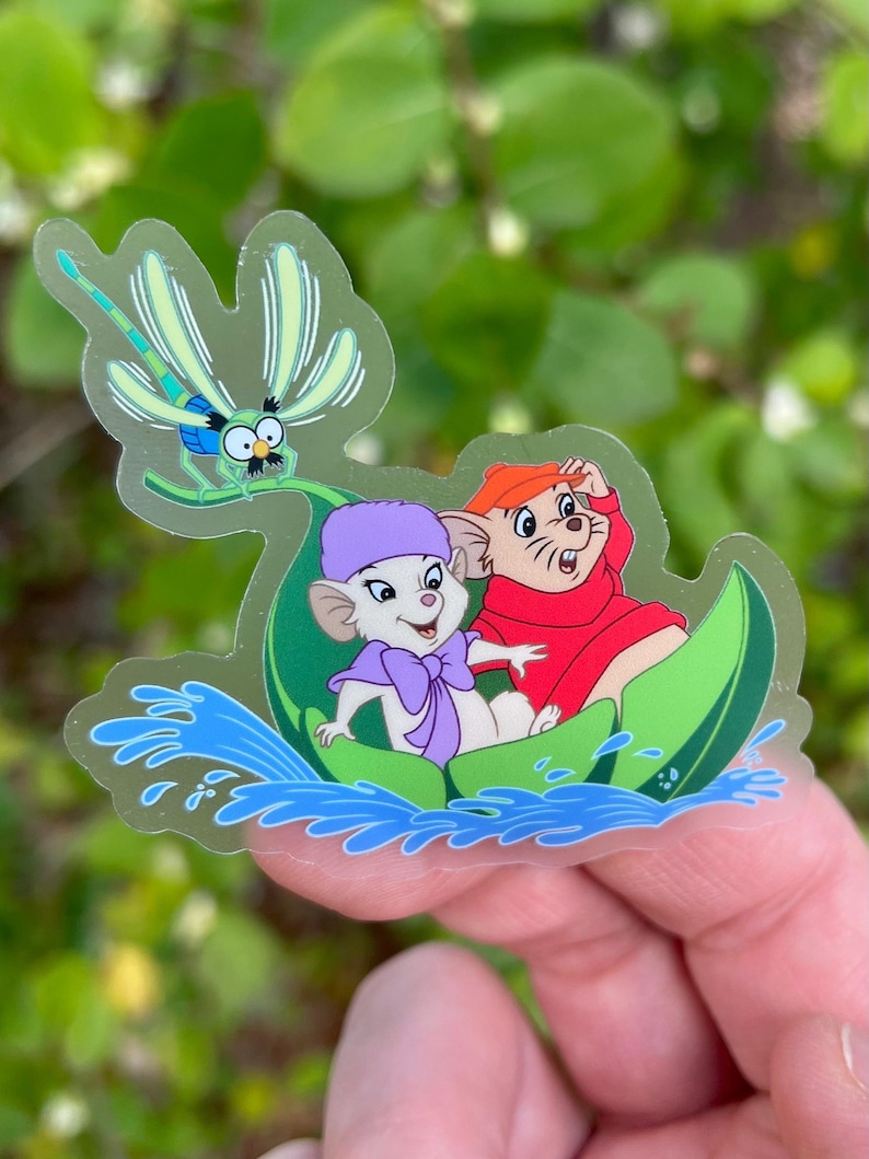 May include: A clear sticker featuring two cartoon mice in a green leaf boat, with a dragonfly flying above them. The mice are wearing hats and the boat is floating on blue water.