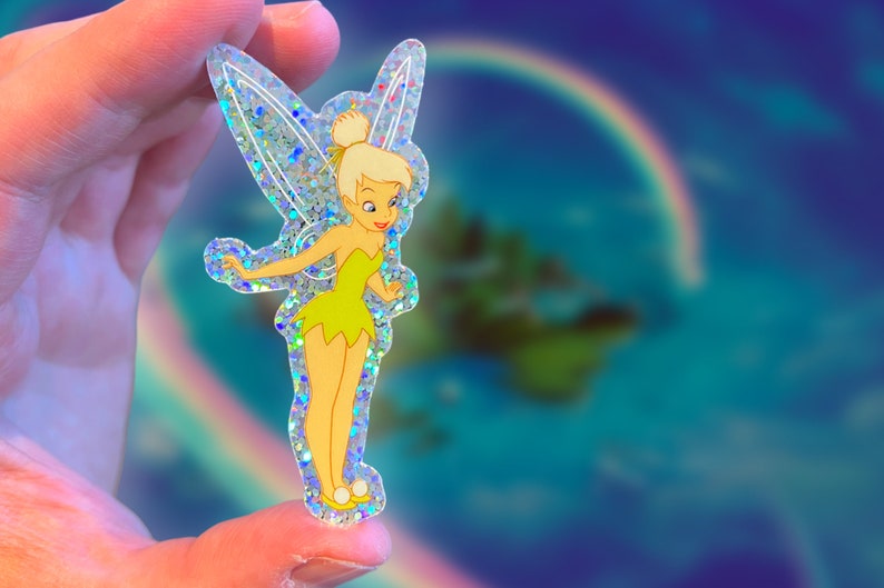 K&ouml;nnte beinhalten: Ein transparenter Aufkleber mit Glitzer-Finish, der Tinkerbell zeigt, eine Fee mit gr&uuml;nen Fl&uuml;geln und einem gr&uuml;nen Kleid, die auf einem blauen und gr&uuml;nen Hintergrund steht.