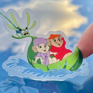 May include: A sticker featuring two cartoon mice riding in a green leaf boat. The mice are wearing hats and are smiling. A dragonfly is flying above them. The background is a blue sky with white clouds.