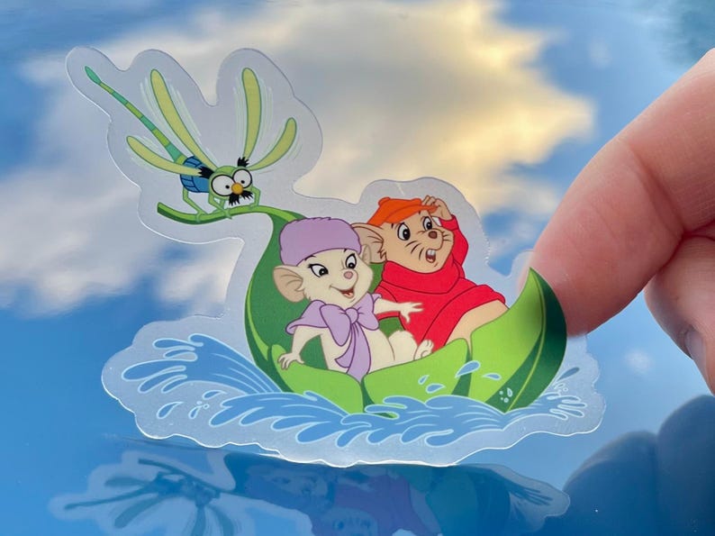 May include: A sticker featuring two cartoon mice riding in a green leaf boat. The mice are wearing hats and are smiling. A dragonfly is flying above them. The background is a blue sky with white clouds.