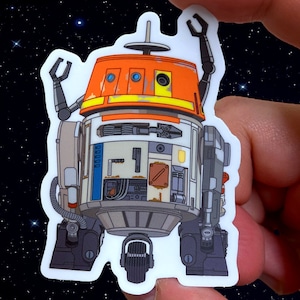 May include: A white, orange, and blue robot sticker with a distressed look. The robot has two arms raised in the air and is standing on two legs. The sticker is on a black background with white stars.