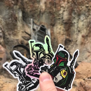 May include: Three Star Wars Boba Fett stickers with holographic details. The stickers are black and white with green and red accents. The stickers are on a white background.