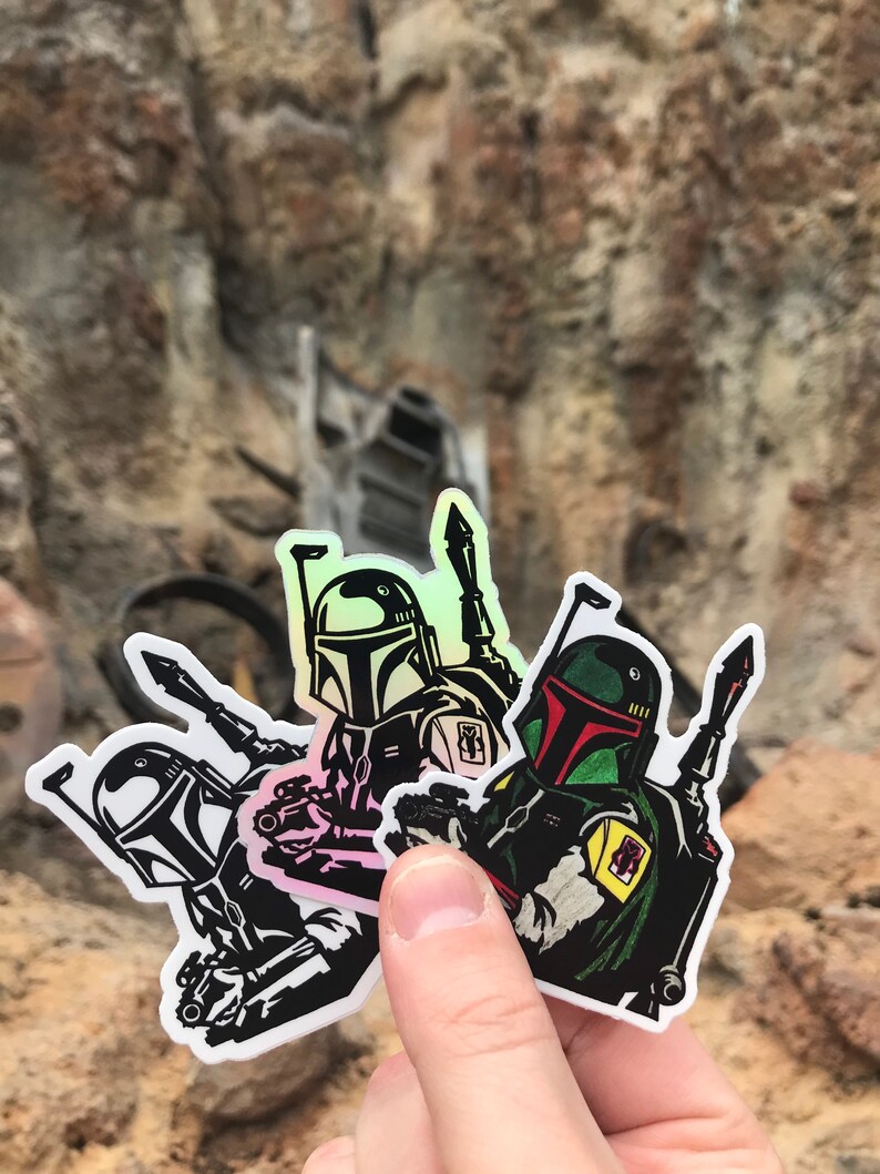 May include: Three Star Wars Boba Fett stickers with holographic details. The stickers are black and white with green and red accents. The stickers are on a white background.