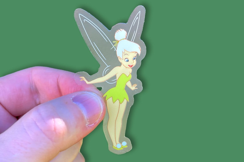 K&ouml;nnte beinhalten: Ein transparenter Aufkleber mit Tinkerbell, einer Fee mit gr&uuml;nen Fl&uuml;geln und einem gr&uuml;nen Kleid. Sie hat blonde Haare und l&auml;chelt.