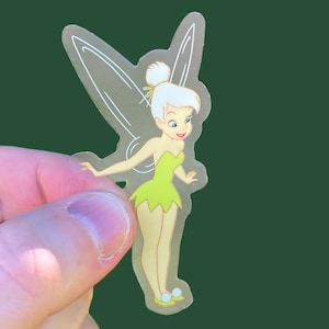 K&ouml;nnte beinhalten: Ein transparenter Aufkleber mit Tinkerbell, einer Fee mit gr&uuml;nen Fl&uuml;geln und einem gr&uuml;nen Kleid. Sie hat blonde Haare und l&auml;chelt.