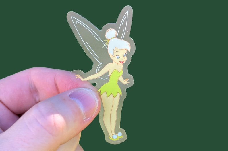 K&ouml;nnte beinhalten: Ein transparenter Aufkleber mit Tinkerbell, einer Fee mit gr&uuml;nen Fl&uuml;geln und einem gr&uuml;nen Kleid. Sie hat blonde Haare und l&auml;chelt.