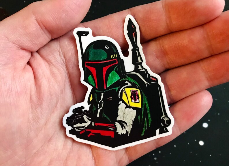 May include: A green and black Boba Fett sticker with a red visor and a yellow shoulder pad. The sticker is on a hand.