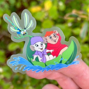 May include: A clear sticker featuring a cartoon mouse wearing a purple hat and a red mouse wearing a red coat. They are both riding in a green leaf boat on a blue water background. A dragonfly is flying above them.