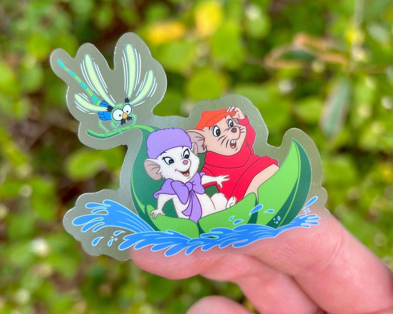 May include: A clear sticker featuring a cartoon mouse wearing a purple hat and a red mouse wearing a red coat. They are both riding in a green leaf boat on a blue water background. A dragonfly is flying above them.