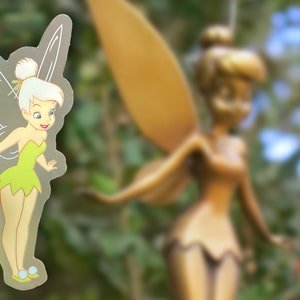 K&ouml;nnte beinhalten: Ein transparenter Aufkleber mit Tinkerbell, einer Fee mit gr&uuml;nen Fl&uuml;geln und einem gr&uuml;nen Kleid, die vor einer gr&ouml;&szlig;eren goldenen Tinkerbell-Statue steht.