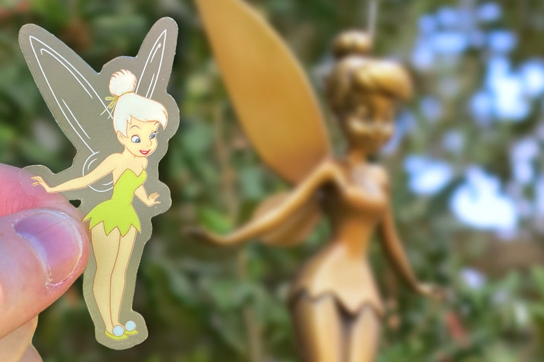 K&ouml;nnte beinhalten: Ein transparenter Aufkleber mit Tinkerbell, einer Fee mit gr&uuml;nen Fl&uuml;geln und einem gr&uuml;nen Kleid, die vor einer gr&ouml;&szlig;eren goldenen Tinkerbell-Statue steht.