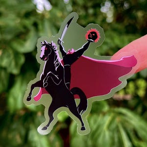 Puede incluir: Una pegatina transparente con un caballo negro encabritado con un jinete sin cabeza sosteniendo una espada y una linterna de calabaza en llamas. El jinete lleva una capa roja. La pegatina tiene un contorno verde.