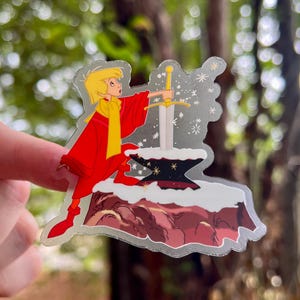 Pode incluir: Um adesivo com um personagem de desenho animado puxando uma espada de uma pedra. O personagem est&aacute; vestindo uma roupa vermelha e amarela e est&aacute; cercado por estrelas brilhantes.