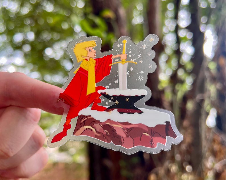 Pode incluir: Um adesivo com um personagem de desenho animado puxando uma espada de uma pedra. O personagem est&aacute; vestindo uma roupa vermelha e amarela e est&aacute; cercado por estrelas brilhantes.