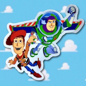 May include: A sticker featuring the characters Woody and Buzz Lightyear from the animated film Toy Story. Woody is wearing a cowboy hat and Buzz is in a spacesuit, both against a blue sky background with white clouds.