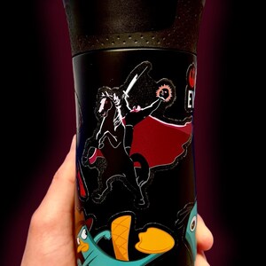 Puede incluir: Taza de viaje negra con tapa negra y pegatinas de personajes de dibujos animados. Las pegatinas incluyen un personaje con espada y capa, y un ornitorrinco turquesa con pico naranja. La taza se sostiene en una mano.