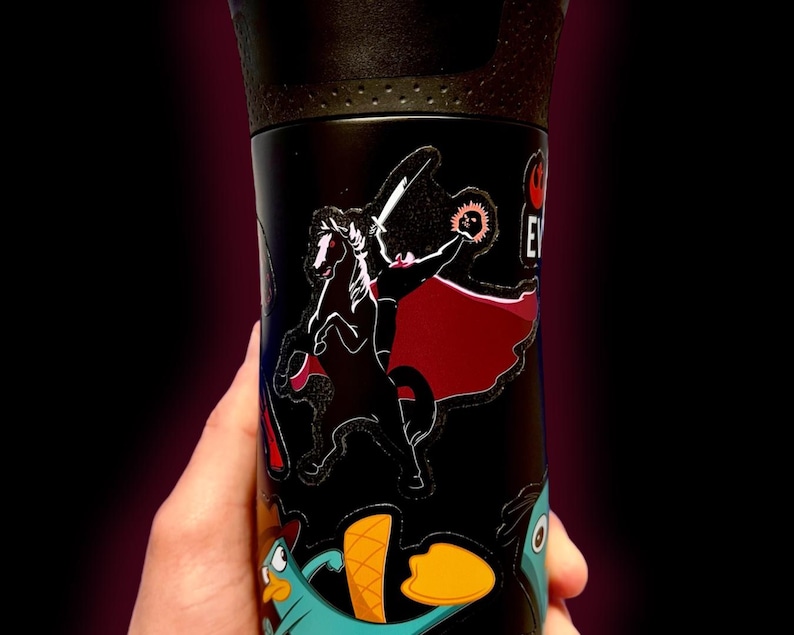 Puede incluir: Taza de viaje negra con tapa negra y pegatinas de personajes de dibujos animados. Las pegatinas incluyen un personaje con espada y capa, y un ornitorrinco turquesa con pico naranja. La taza se sostiene en una mano.
